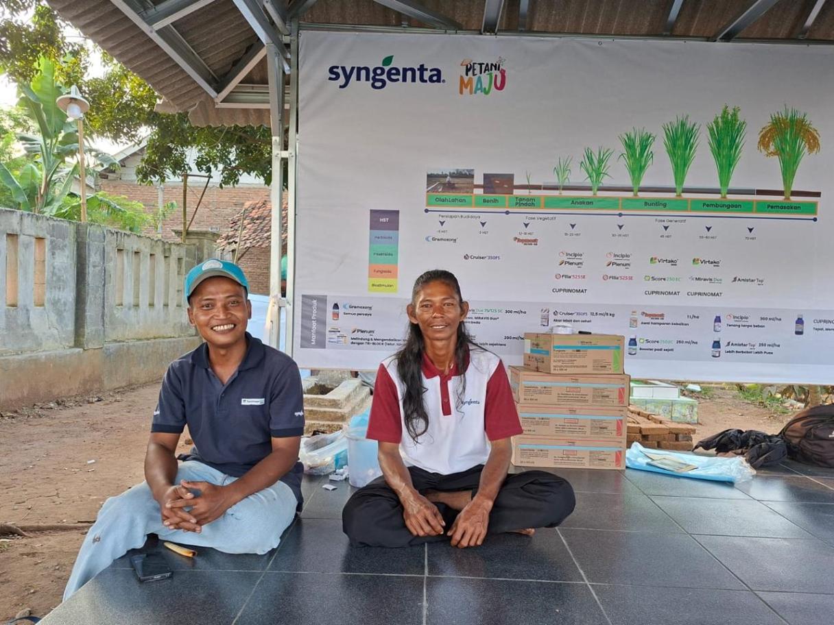 Dua petani duduk di lantai, di belakangnya papan grafik dan kotak-kotak.