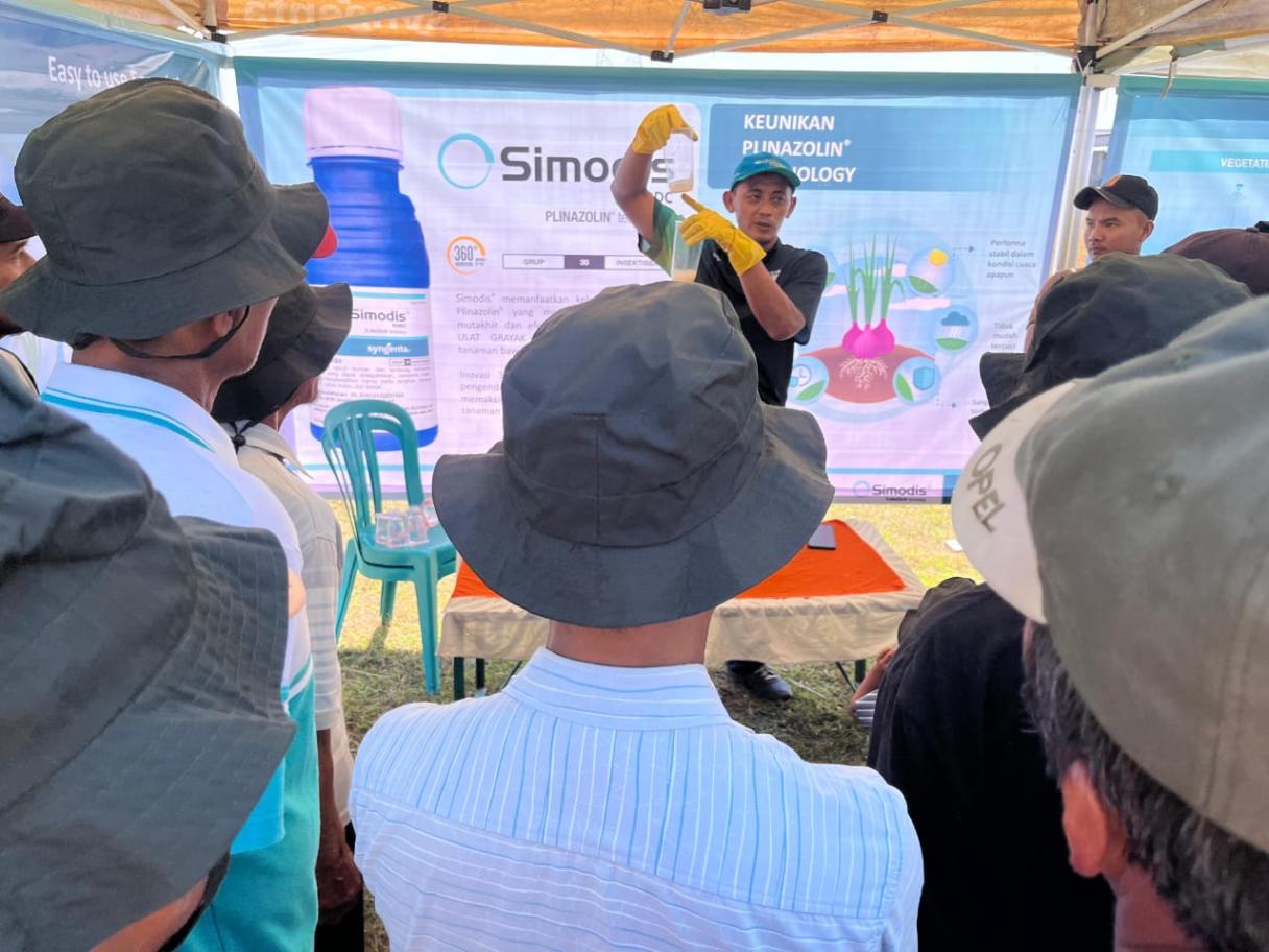 Sekelompok orang ber-topi melihat presentasi di stan pameran luar ruangan.